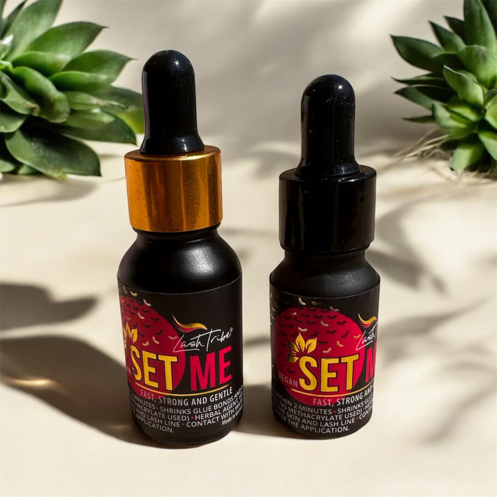 Two small black dropper bottles labeled "SET ME" stand against a light background, with two green succulent plants in the backdrop. One bottle with a gold cap holds Vegan Setting Liquid, while the other with a black cap contains Lash Tribe's Superbonder for Eyelash Extensions.