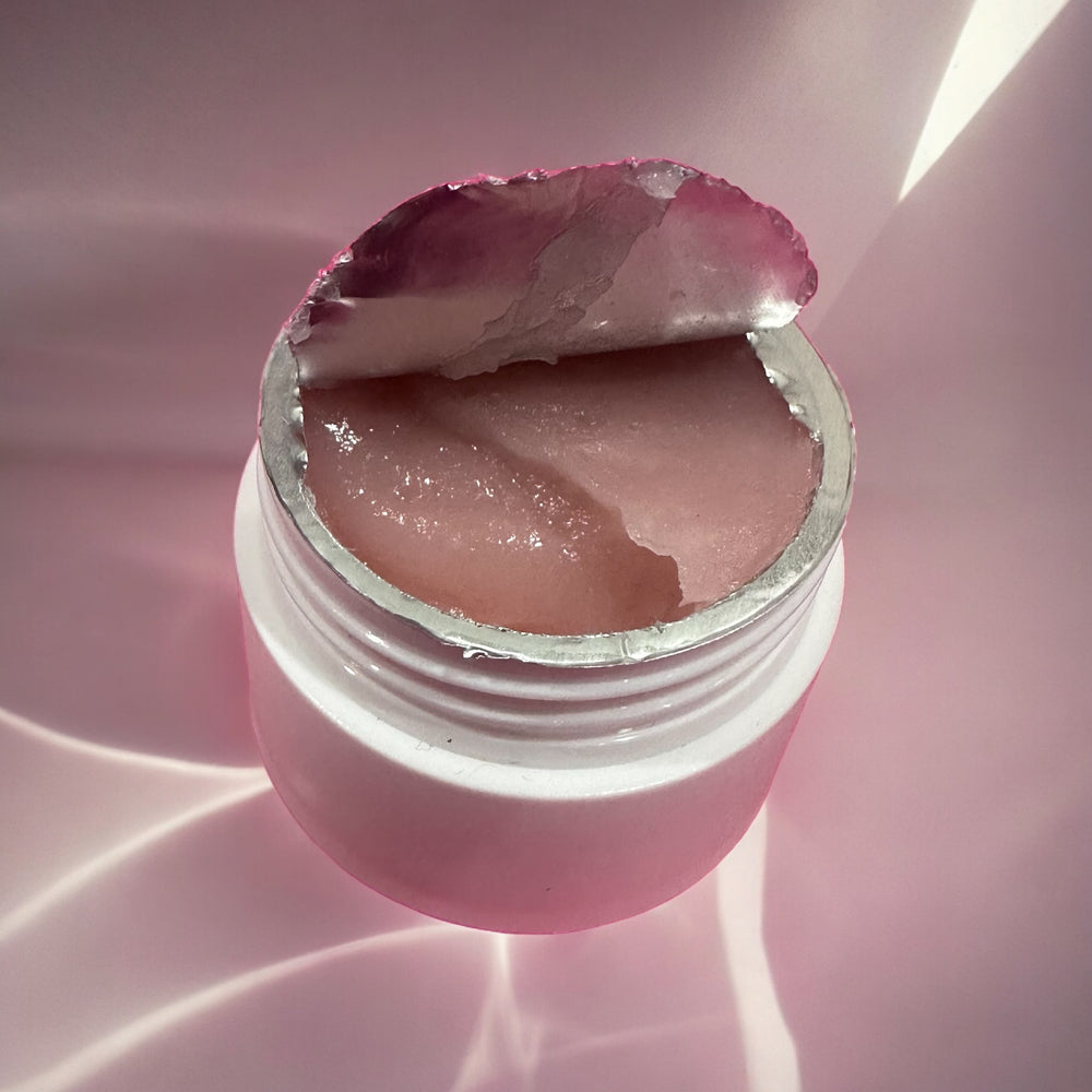 An open white jar with a partially peeled metallic seal revealing a pink, gel-like substance inside, perfect for professional use as an eyelash extensions remover, set against a pink background. The product is Lash Tribe® Cream Remover by Lash Tribe.