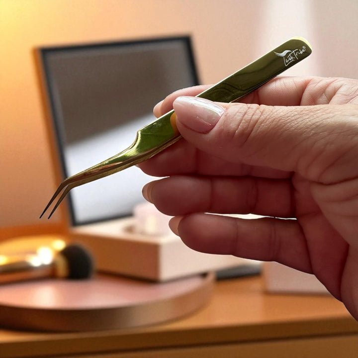 A hand holds the Lash Tribe Classic Duo - Lash Tweezers | Pickup and Isolation near a makeup table with a mirror, brush, and other cosmetics in the background.