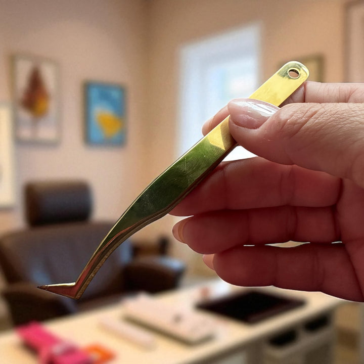 A hand holds the Lash Tribe Classic Duo - Lash Tweezers | Pickup and Isolation (gold-colored) in front of a blurred office background with a desk and chair.