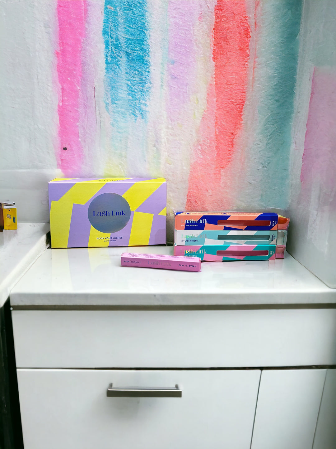 Boxes of Lash Link DIY Eyelash Extension Kits are arranged on a white cabinet, perfect for your Eyelash Extension Home Studio. The wall behind them is painted with colorful stripes in shades of pink, blue, and orange.