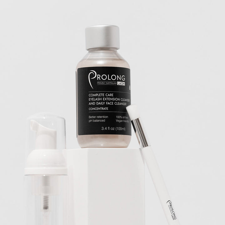 A bottle of PROLONG LASH | EYELASH EXTENSION CLEANSER CONCENTRATE 100ml by Lash Tribe and a brush on top of a white surface.