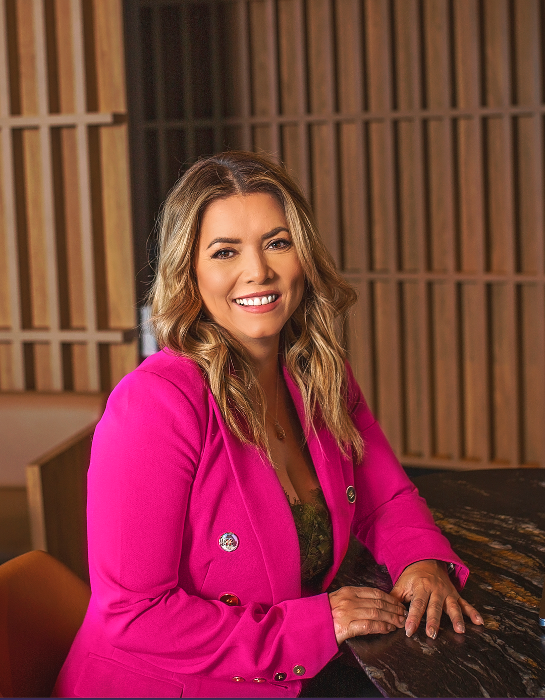 Julia Mann Lash Trainer sitting at a table and smiling into the camera wearing a bright pink blazer