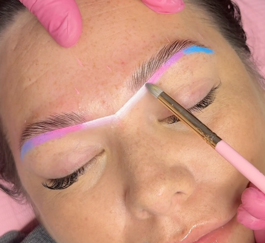 A close-up shot of a face during an eyebrow treatment features a gloved hand holding the skin while vibrant colors from the Lash Tribe Infinity Hybrid Metallic Mapping Stack - 6pk are expertly brushed above the brow, highlighting exquisite artistry.