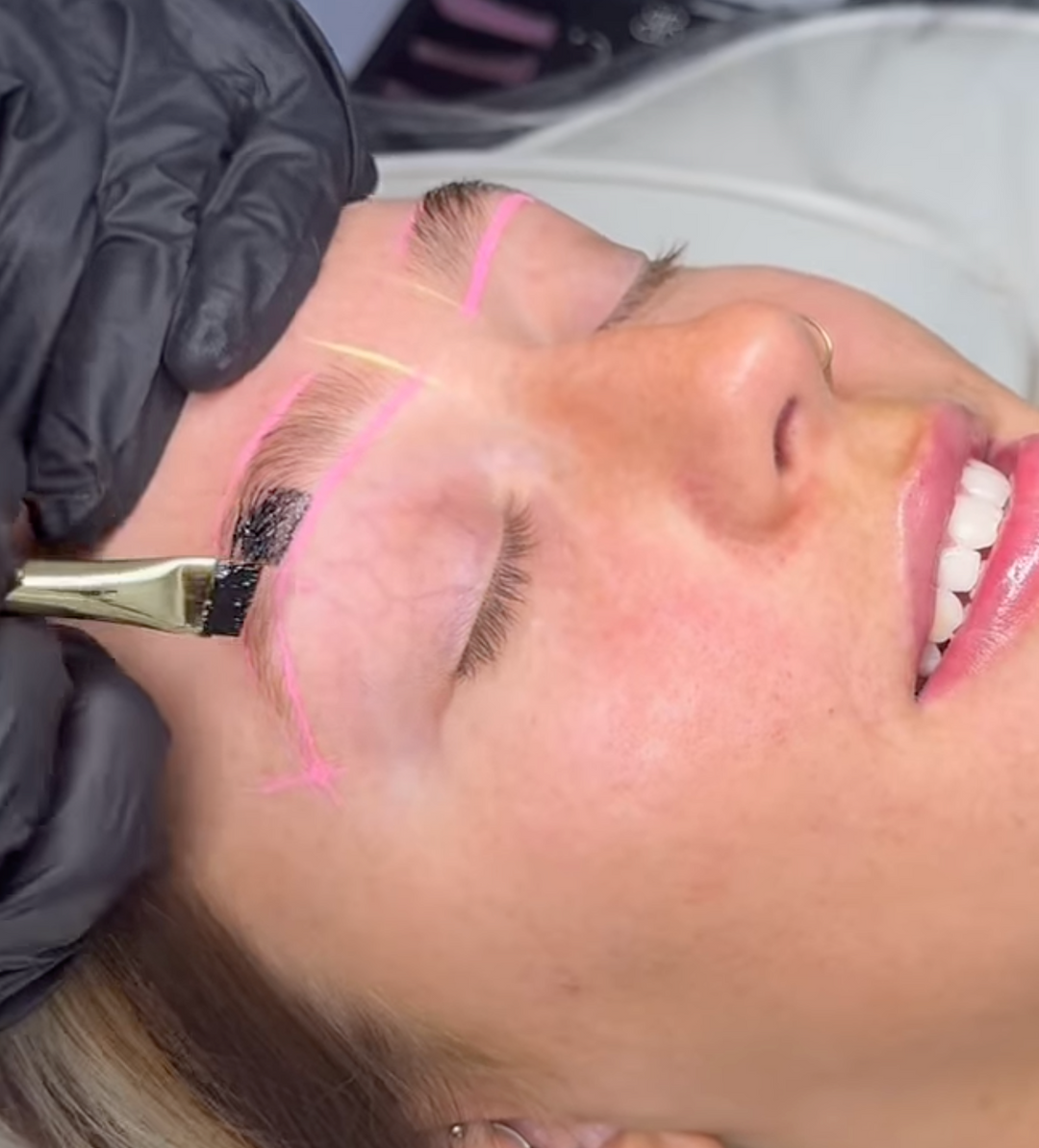 A person with closed eyes enjoys brow artistry as their eyebrows are tinted. A black substance, enhanced by vibrant mapping paste colors, is applied with a brush, while pink guidelines from the Lash Tribe Infinity Hybrid Metallic Mapping Stack - 6pk are visible.