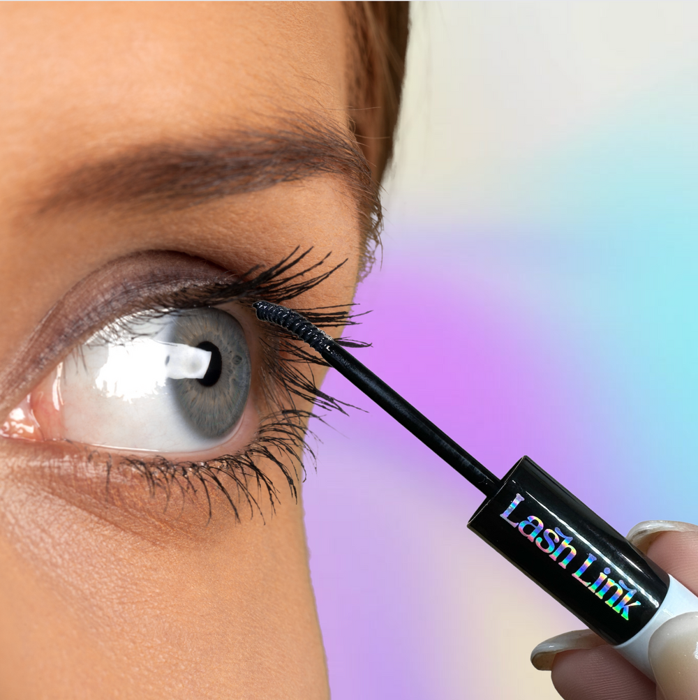 Close-up of a person expertly using the Lash Link Applicator | Lash Segment Tweezer by Lash Link for flawless mascara on upper eyelashes. A soft pastel gradient in the background enhances the scene's elegance.