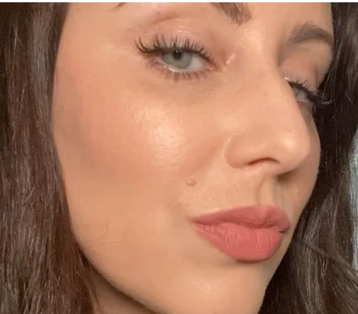 In a home studio, someone with long dark hair and light eyes looks slightly away from the camera, enhanced by pink lipstick and a cheek beauty mark. The elegance of their gaze is further highlighted by subtle extensions using the Lash Link DIY Eyelash Extension Kit.