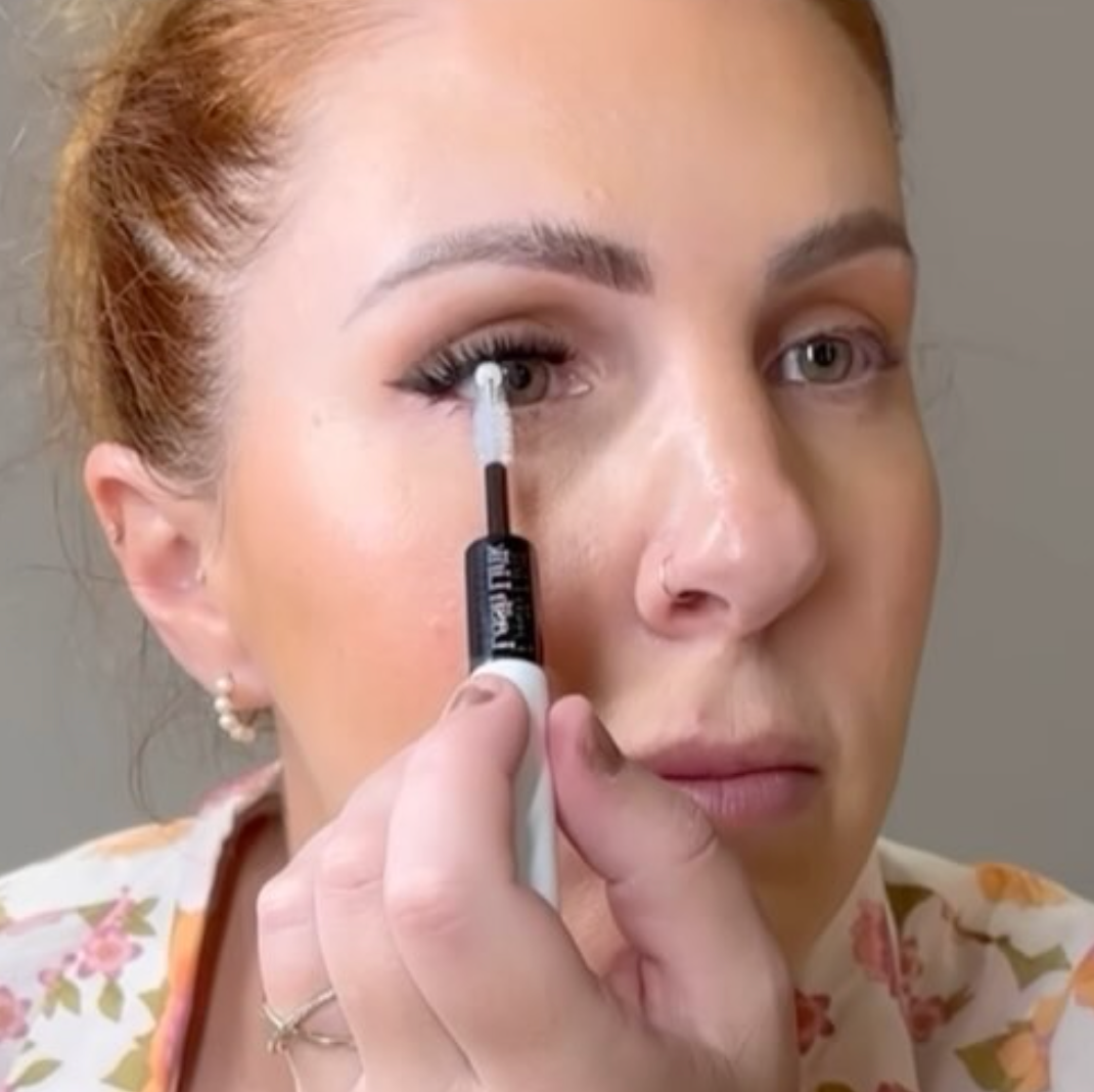 Wearing a floral top and small hoop earrings, the individual expertly applies mascara to her upper lashes using the Lash Link DIY Eyelash Extension Kit.