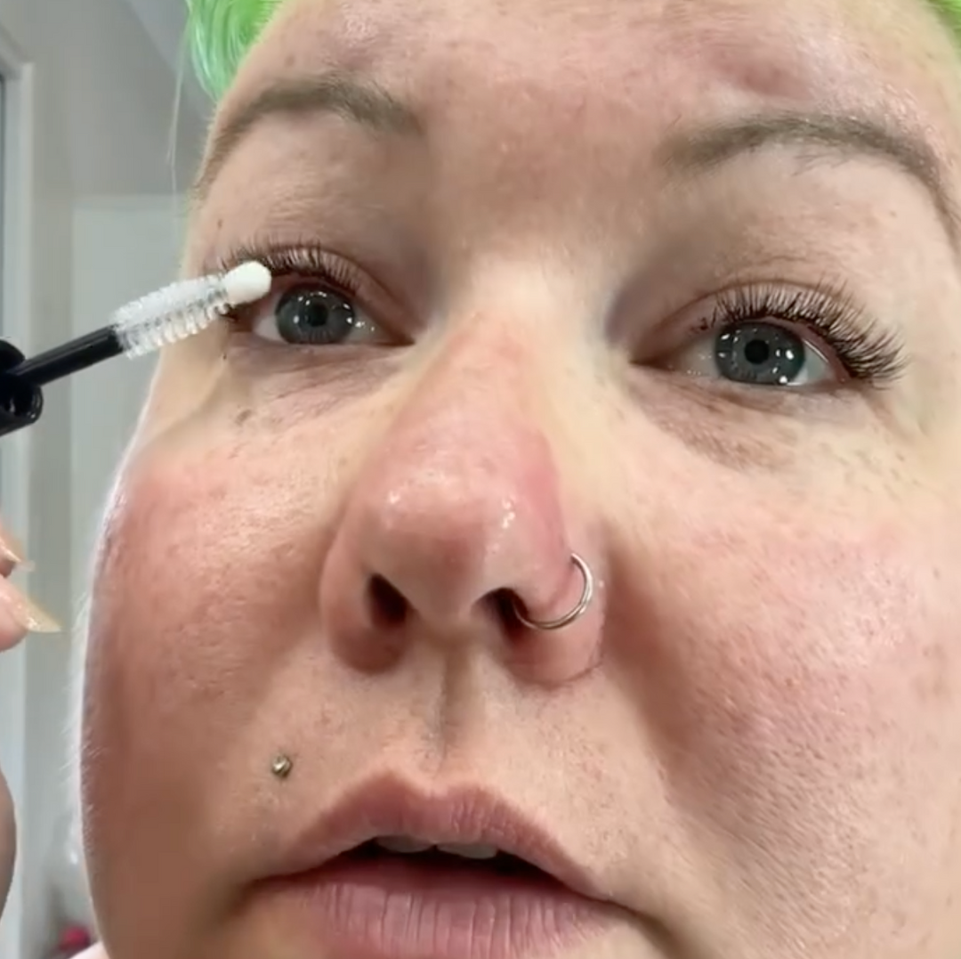 In a cozy home studio, a person with green hair carefully applies mascara to their eyelashes using the brush from their Lash Link DIY Eyelash Extension Kit.
