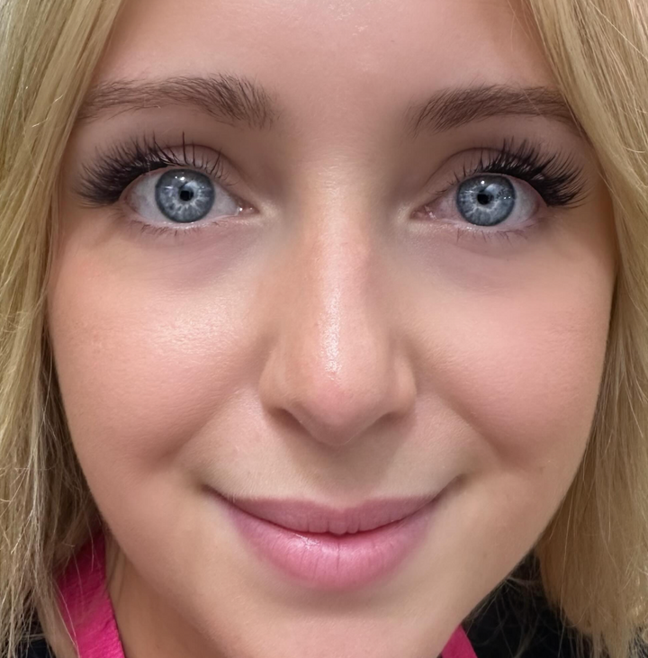 A person with blue eyes and light blonde hair smiles in a cozy home studio while showcasing their natural beauty without needing the Lash Link DIY Eyelash Extension Kit. They're wearing a pink top.