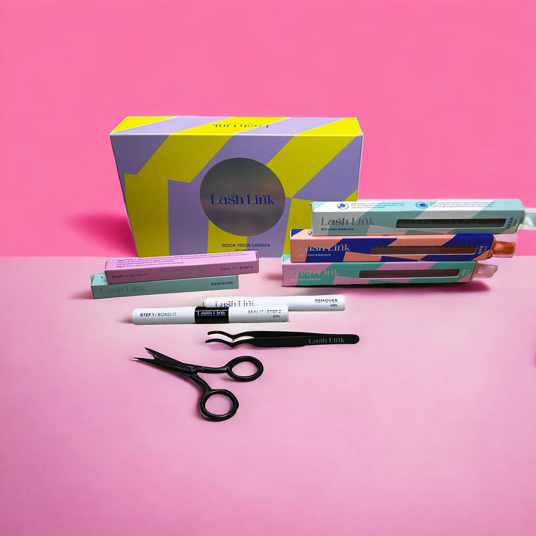 On a pink background, the Lash Link DIY Eyelash Extension Kit showcases scissors, tweezers, and assorted pastel-colored labeled product boxes and tubes, ideal for trying different lash styles.