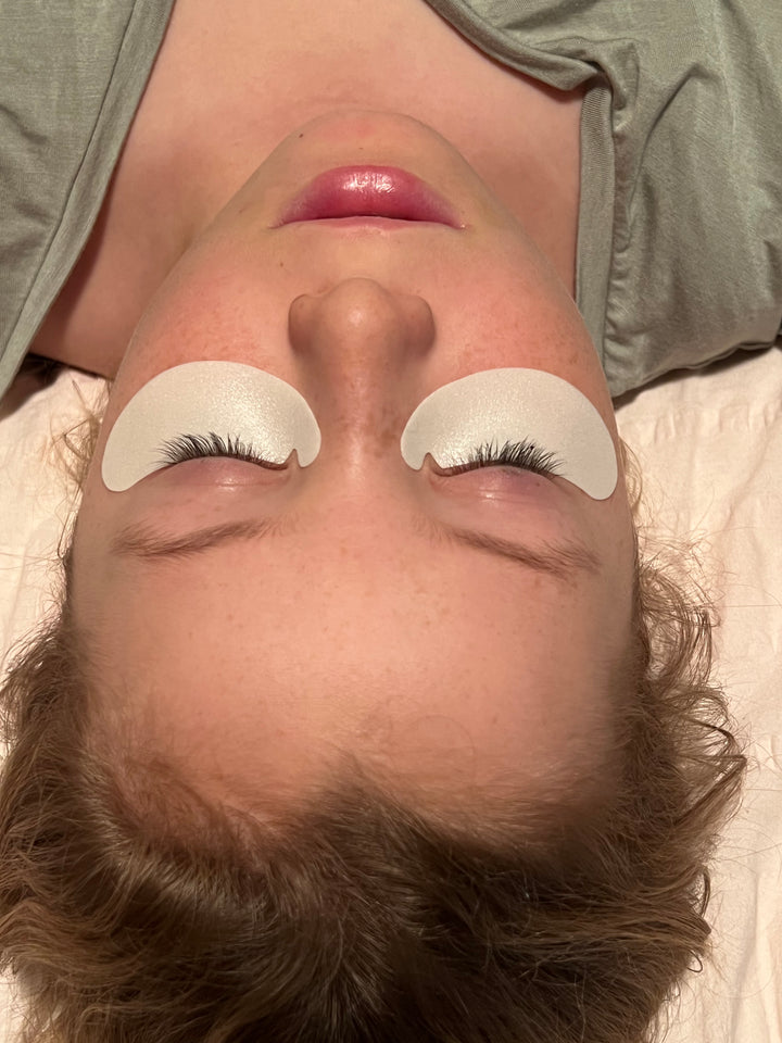 A woman laying on a bed with her eyes closed, wearing Lash Tribe Microfoam Eye Pads.