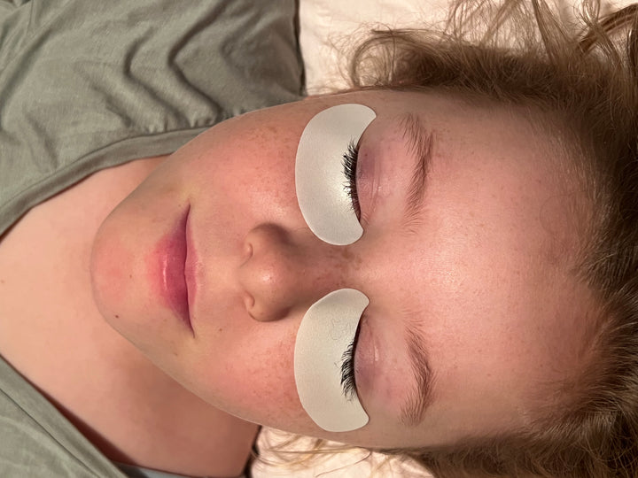 A woman laying on a bed with a pair of Lash Tribe Microfoam Eye Pads.